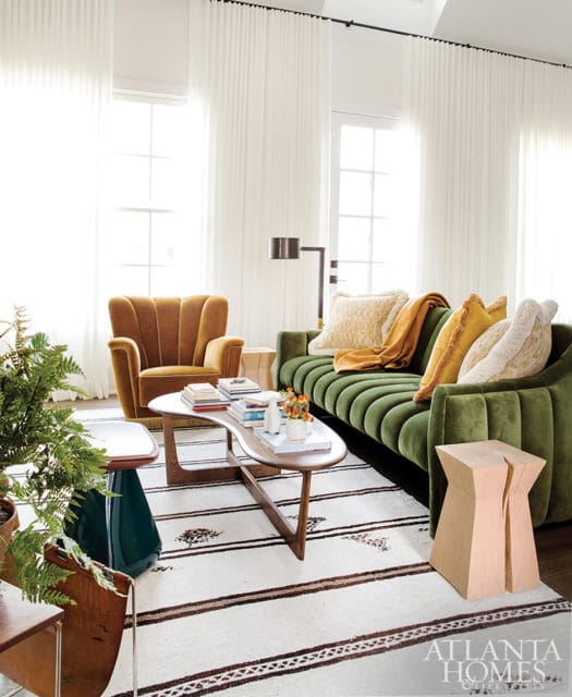 Living room with a green channel-tufted sofa, rust velvet armchair, organic wood coffee table, and a striped rug. White floor-to-ceiling curtains frame large windows.