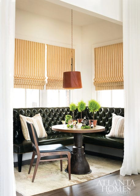 Dining nook with a dark green tufted leather banquette, round wood table with potted plants and bottles, and striped roman shades.