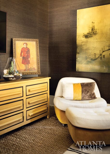Dark textured wallpaper adorns the walls of a room featuring a yellow dresser with black trim, a terrarium, a white armchair with a striped pillow, and a large abstract painting of a ship.