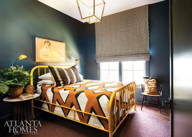 Bedroom with dark blue walls, a yellow metal bed with an orange and white geometric quilt, a patterned Roman shade, and a painting of a reclining figure above the bed.
