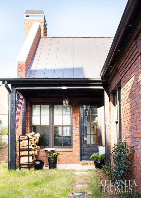 Brick house with a gray metal roof, black trim, and a covered porch with an American flag and stacked firewood.