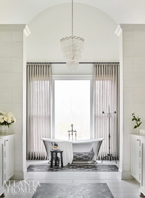 White bathroom with a freestanding silver bathtub, crystal chandelier, floor-to-ceiling marble tile, and patterned floor tiles.