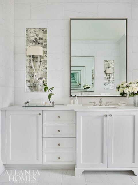 White bathroom vanity with marble tile walls, a large framed mirror, and decorative sconces.