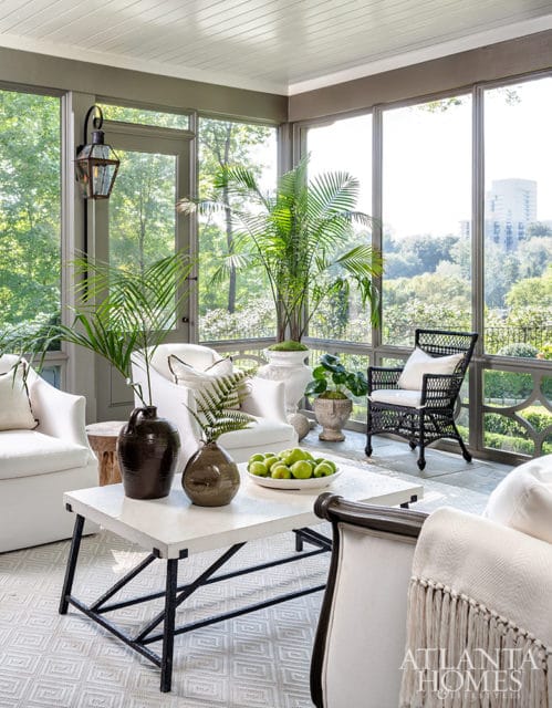 Screened-in porch with white upholstered chairs, a white coffee table with dark vases and green apples, and a black wicker chair. Large windows overlook a lush green landscape.