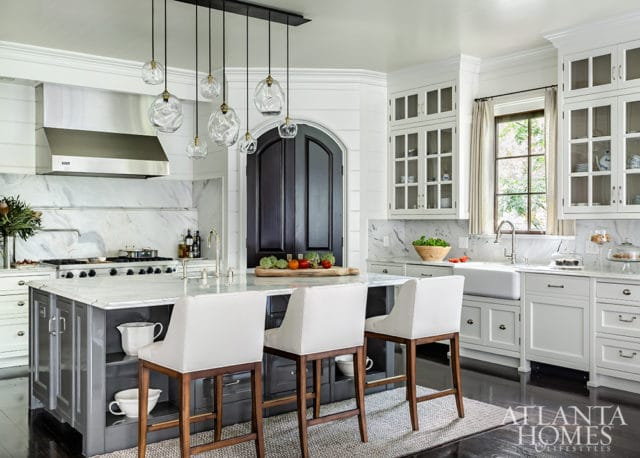 White kitchen with marble countertops, a large island with white bar stools, stainless steel range, and glass pendant lights over the island.