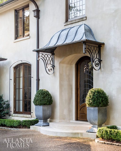 Exterior of a home with stucco walls, arched windows, a metal awning over a wooden door, and two large planters with boxwood shrubs.