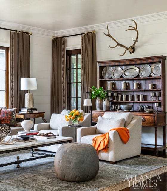 Living room with light gray armchairs, a marble coffee table, a dark wood hutch filled with pewter dishes, and elk antlers mounted on the wall.