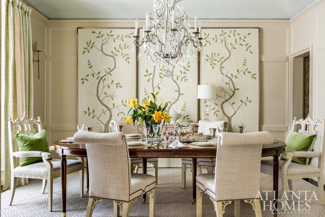 Dining room with a dark wood oval table set for a meal, surrounded by upholstered chairs and ornate white chairs with green cushions. A crystal chandelier hangs above, and three panels of chinoiserie wallpaper featuring trees and birds adorn the wall.