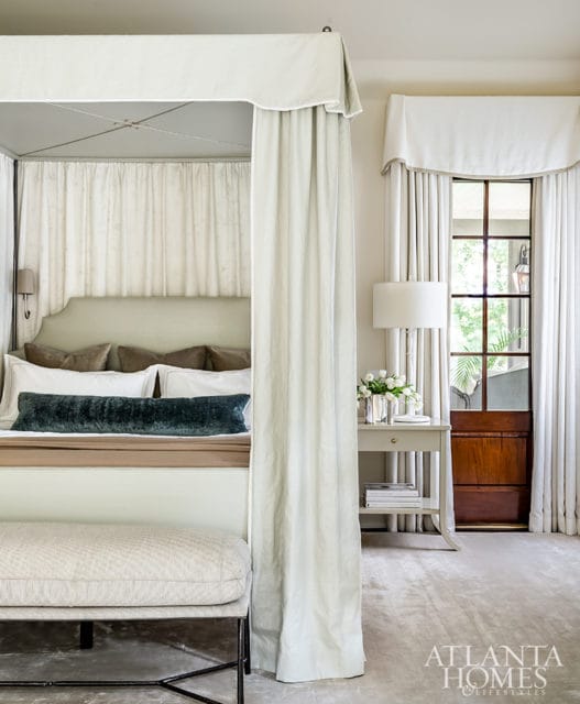 Bedroom with a four-poster bed draped in light fabric, upholstered headboard, and plush pillows. A bench sits at the foot of the bed. A side table with a lamp and flowers is next to a wooden door and window.