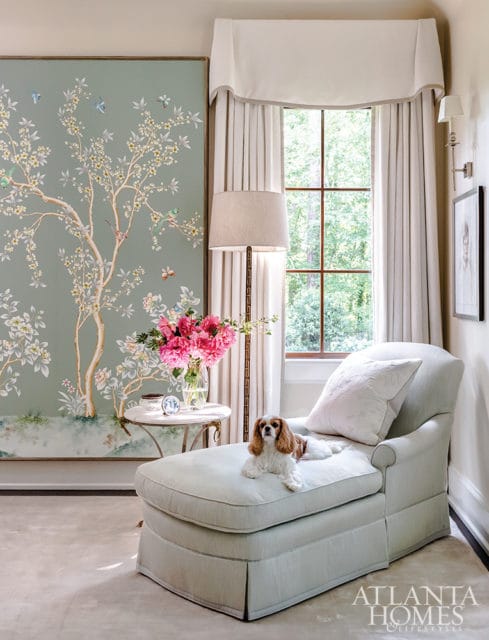 Light blue chaise lounge with a Cavalier King Charles Spaniel dog resting on it, next to a round side table with a vase of pink flowers. A large chinoiserie mural adorns the wall.