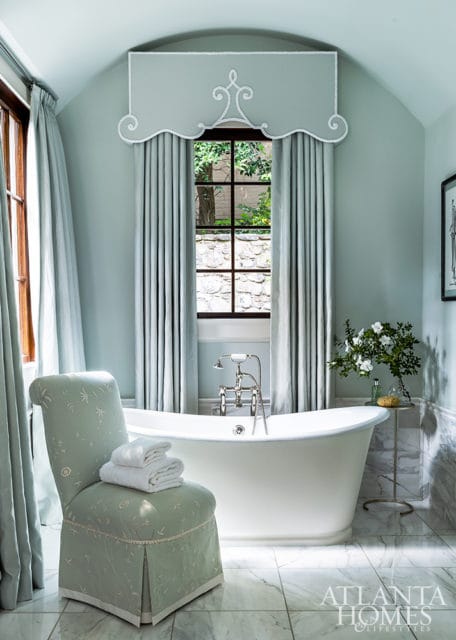 Light blue bathroom with a freestanding white bathtub, a patterned upholstered chair with stacked white towels, and marble tile flooring.