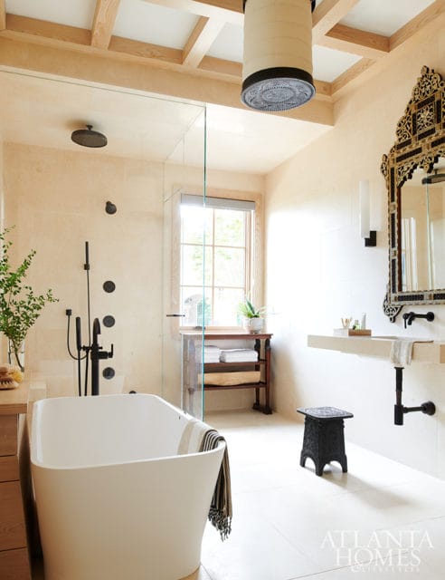 Cream tiled bathroom with a freestanding white tub, glass-enclosed shower with black fixtures, stone vanity, and ornate black mirror above the sink.