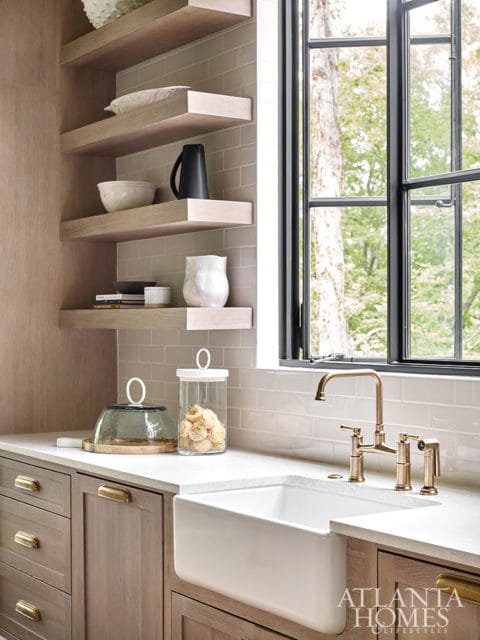 Kitchen with light wood cabinetry, white subway tile backsplash, farmhouse sink, and brass bridge faucet. Open shelving displays decorative bowls and vases.