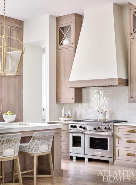 Light wood kitchen with stainless steel range, white subway tile backsplash, and a large white range hood. Two woven bar stools sit at a stone-topped island.