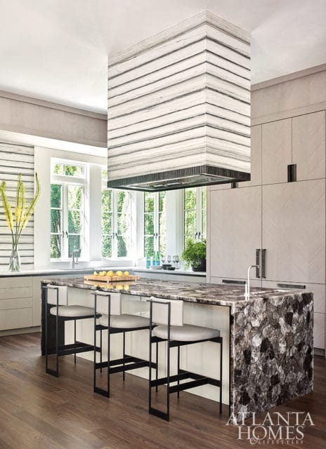 Modern kitchen with a large island featuring a dark, multi-faceted stone countertop and base, surrounded by light wood cabinetry and a striped marble range hood.