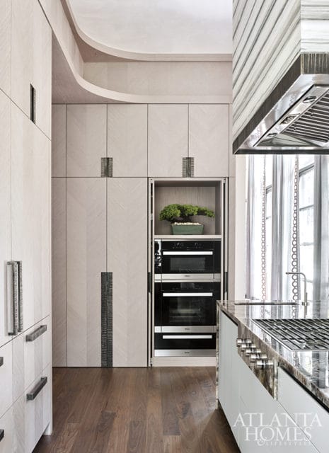 Modern kitchen with light wood cabinetry in a herringbone pattern, stacked ovens, and a marble countertop with a gas range.