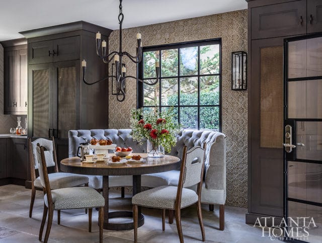 Dark wood kitchen with a round dining table, upholstered banquette seating, and patterned wallpaper. A large window overlooks greenery.
