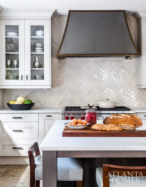White kitchen with glass-front cabinetry, herringbone tile backsplash, and a large metal range hood. A marble island in the foreground holds a cutting board with bread and a bowl of eggs.