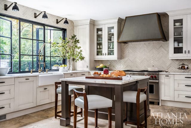 Cream kitchen with marble island, dark wood base, white leather stools, herringbone tile backsplash, and large black-framed window overlooking greenery.