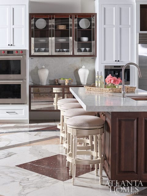 White kitchen with glass-front cabinetry, double ovens, marble countertops, and bar stools at a dark wood island.