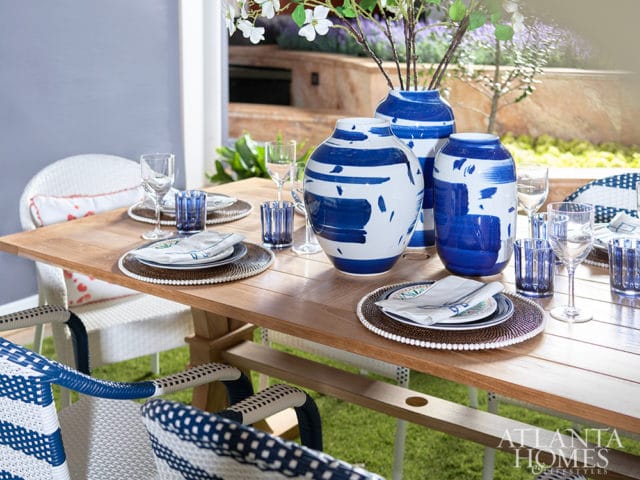Wooden dining table set with blue and white patterned chairs, blue glass tumblers, white plates, and blue and white patterned vases filled with flowers.
