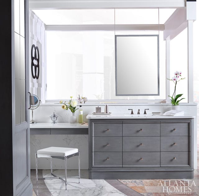 Gray vanity with nine drawers and marble countertop, white upholstered stool, and decorative wall art in a bright bathroom.