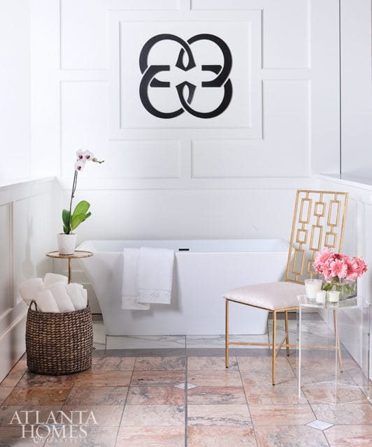 White freestanding bathtub with towels draped over the side, artwork on the wall, a woven basket of towels, and a gold-accented stool next to an acrylic side table with flowers.