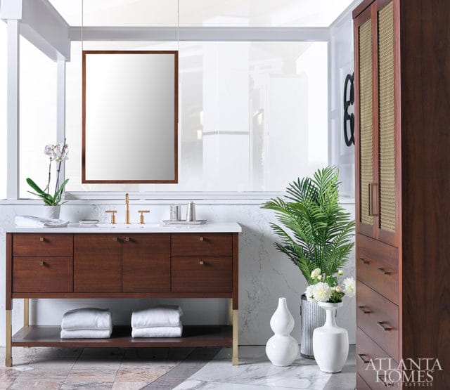 Modern bathroom with a dark wood vanity featuring brass hardware and a marble countertop. A large mirror hangs above, and a tall wood cabinet with cane detailing stands to the right.