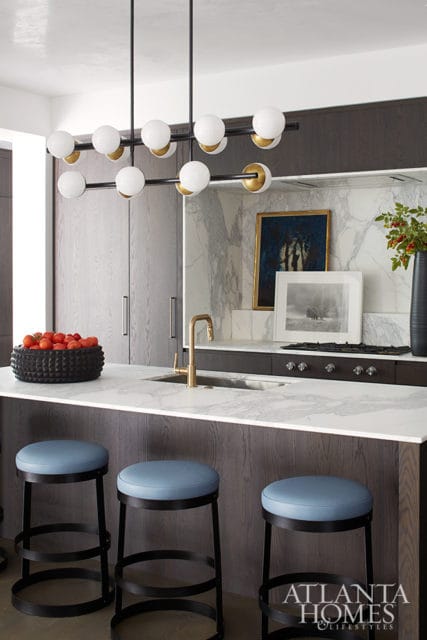 Modern kitchen island with white marble countertop, gold faucet, and blue upholstered stools. Dark wood cabinetry and marble backsplash with artwork.