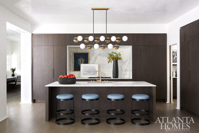 Dark wood kitchen with white marble backsplash, island with blue stools, and a modern brass chandelier. Atlanta Homes & Lifestyles logo in the lower right corner.
