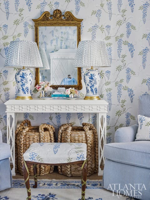 Blue and white floral wallpaper adorns this room, featuring a white console table with two blue and white porcelain lamps and a gold ornate mirror. Below the table are woven baskets and a floral upholstered stool.