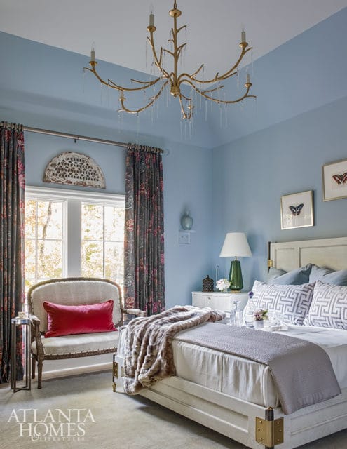 Light blue bedroom with a white bed, patterned pillows, a fur throw, and a vintage armchair with a red pillow. A gold chandelier hangs from the ceiling.