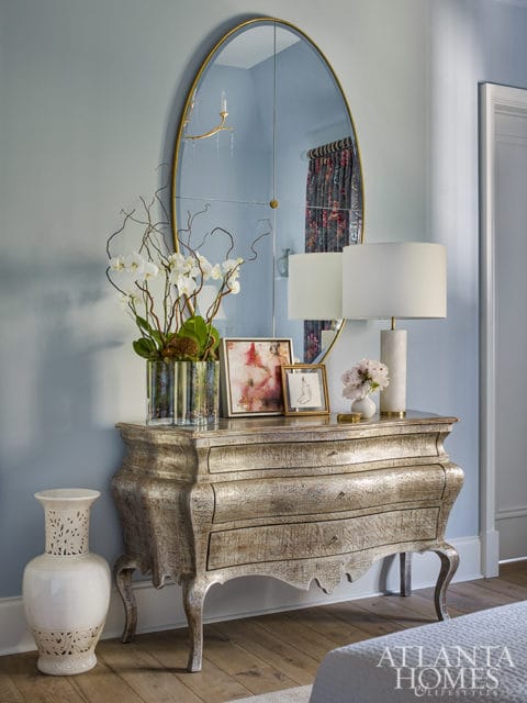 Light blue walls with an ornate antique dresser topped with orchids, framed art, and lamps. A tall white vase sits to the left.