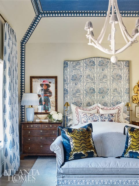 Bedroom with blue and white floral headboard, matching curtains, and a white branch-like chandelier. A dark wood dresser holds a lamp and framed artwork.