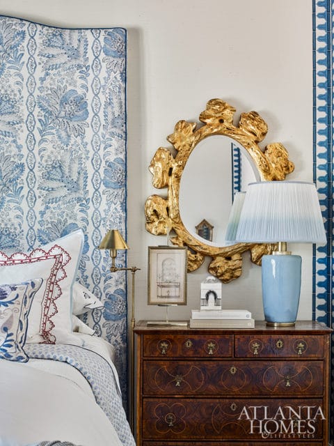 Bedroom with blue and white floral upholstered headboard, antique wooden dresser, gold ornate mirror, and a pale blue lamp.