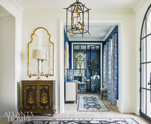 Hallway with a black and gold ornate cabinet topped with a lamp, a gold ornate mirror, and a black metal lantern hanging from the ceiling. Blue walls and patterned curtains are visible through an open doorway.