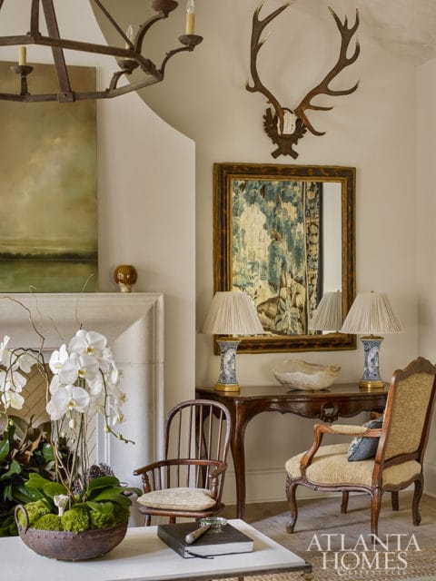 Living room with a stone fireplace, large metal chandelier, and an elk antler mount above a mirror with a tapestry.