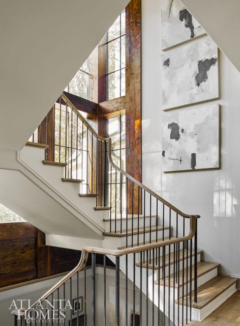 Curved staircase with white risers, natural wood treads, and black metal balusters. Large windows with exposed wooden beams overlook the exterior. Three abstract paintings hang on the wall.