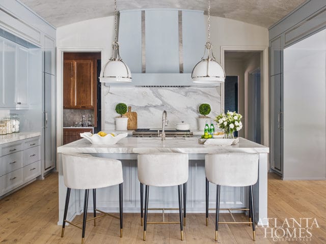 Light blue kitchen with marble island, white velvet bar stools, marble backsplash, and light wood floors.