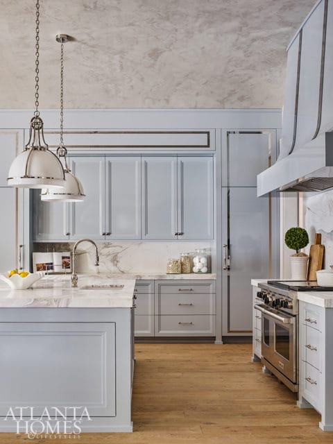 Light blue kitchen with marble countertops, stainless steel range, and pendant lights hanging over island.
