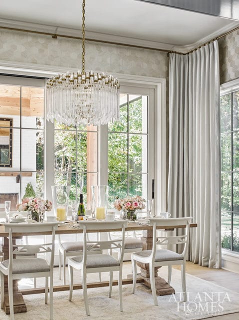 Dining room with a wooden table set for a meal, white chairs, and a crystal chandelier. Large windows overlook a garden.