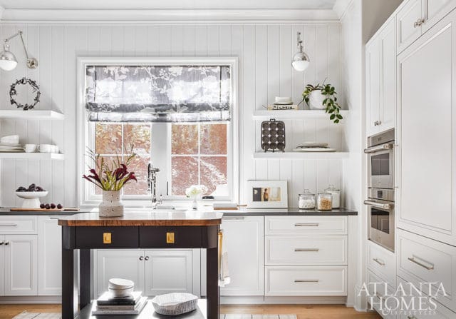 White kitchen with beadboard walls, a dark wood island with brass hardware, stainless steel double ovens, and a floral Roman shade over a window.