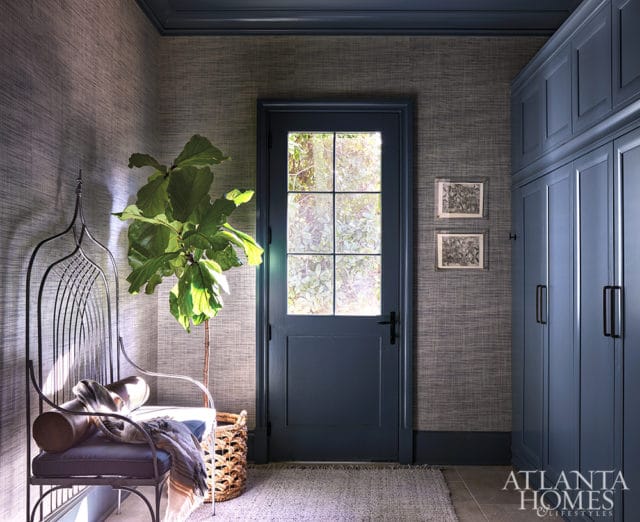 Hallway with textured grey wallpaper, a dark blue door with a glass pane, a metal bench with cushions and a throw, a fiddle leaf fig plant in a basket, and blue built-in cabinets.