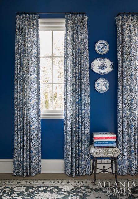 Blue room with patterned curtains, decorative plates on the wall, a stack of books on a stool, and a patterned rug.