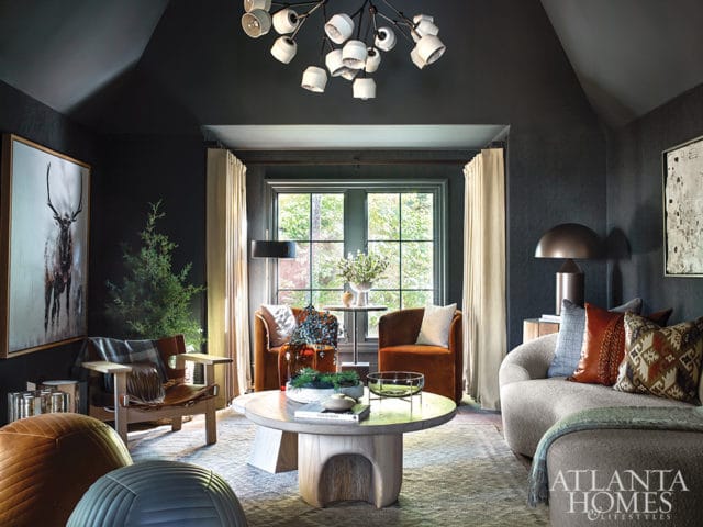 Dark moody living room with vaulted ceiling, featuring a large painting of a deer, orange velvet chairs, a light wood coffee table, and a sectional sofa with patterned pillows.