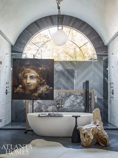 Modern bathroom with freestanding white tub, arched window, herringbone tile shower walls, and artwork of a woman's face