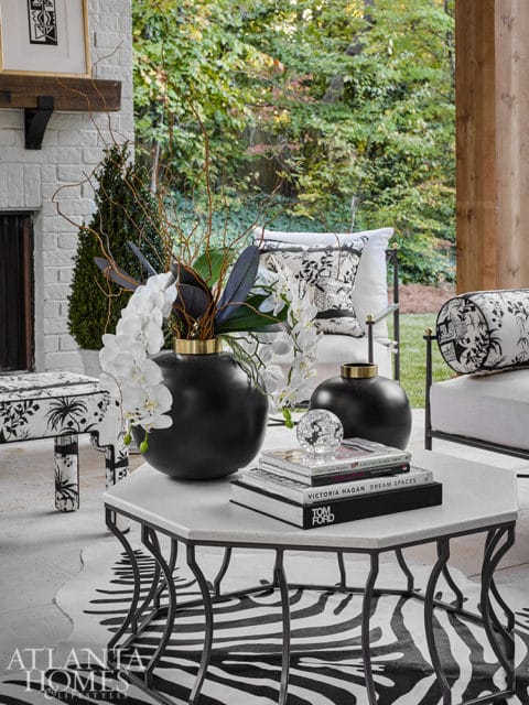 Outdoor living space with white brick fireplace, black and white patterned furniture, and a hexagonal coffee table with books and floral arrangements.