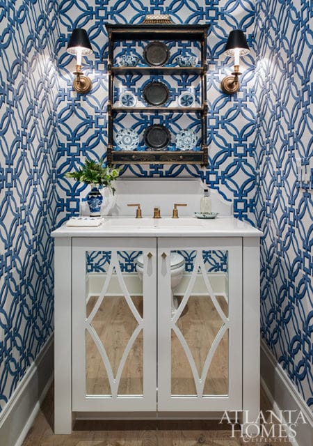 Powder room with blue and white geometric wallpaper, white vanity with mirrored doors, brass fixtures, and a black open shelf displaying decorative plates.