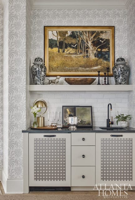 Bar area with gray leaf-patterned wallpaper, white subway tile backsplash, and a dark countertop. Features include a painting, decorative urns, a gold lamp, champagne flutes, a silver ice bucket with champagne, and a black faucet.