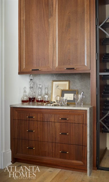 Walnut cabinetry with brass hardware, marble countertop, crystal decanters filled with red liquid, framed artwork, and glassware.